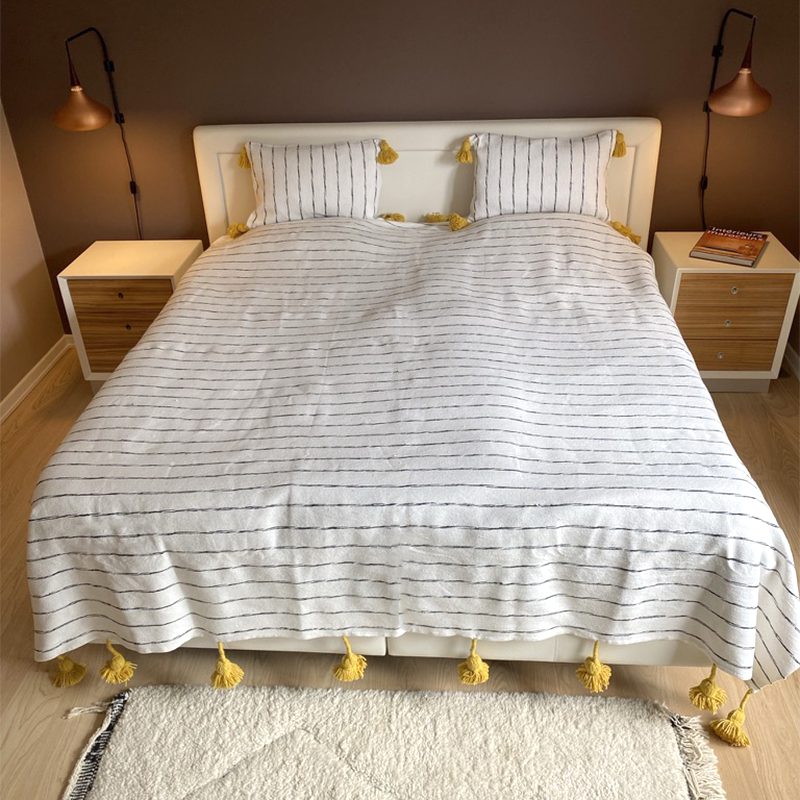 Sengetaeppe ASFAR 1.jpg Witte Marokkaanse handgeweven sprei met zwarte strepen en gele pompons, op een bed met bijpassende kussens