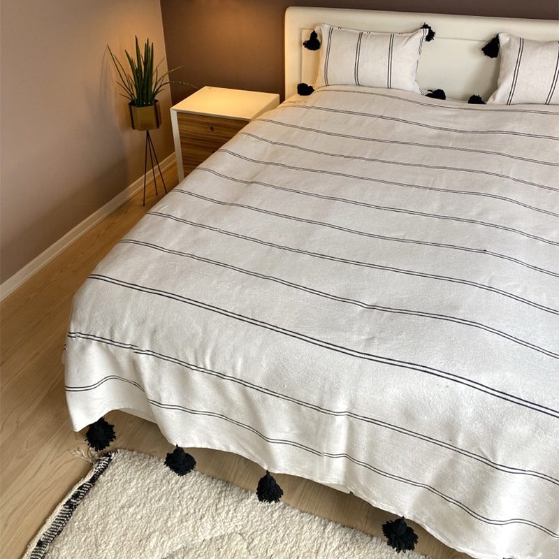 Sengetaeppe ASWAD 1.jpg Witte Marokkaanse handgeweven sprei met zwarte strepen en zwarte pompons op bed met bijpassende kussens