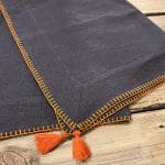 Twee antracietgrijze Marokkaanse handgeborduurde placemats met oranje rand en oranje pompons, op elkaar gevouwen, dicht
