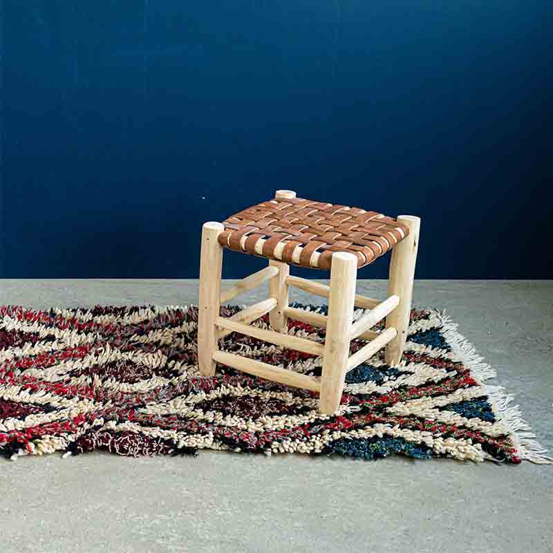 Tibladin Cosy Laine LS 1.jpg Tabouret marocain en bois fait main avec siège en cuir tressé, posé sur un tapis avec un mur bleu derrière