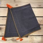 Twee antracietgrijze Marokkaanse handgeborduurde placemats met oranje rand en oranje pompons, op elkaar gevouwen