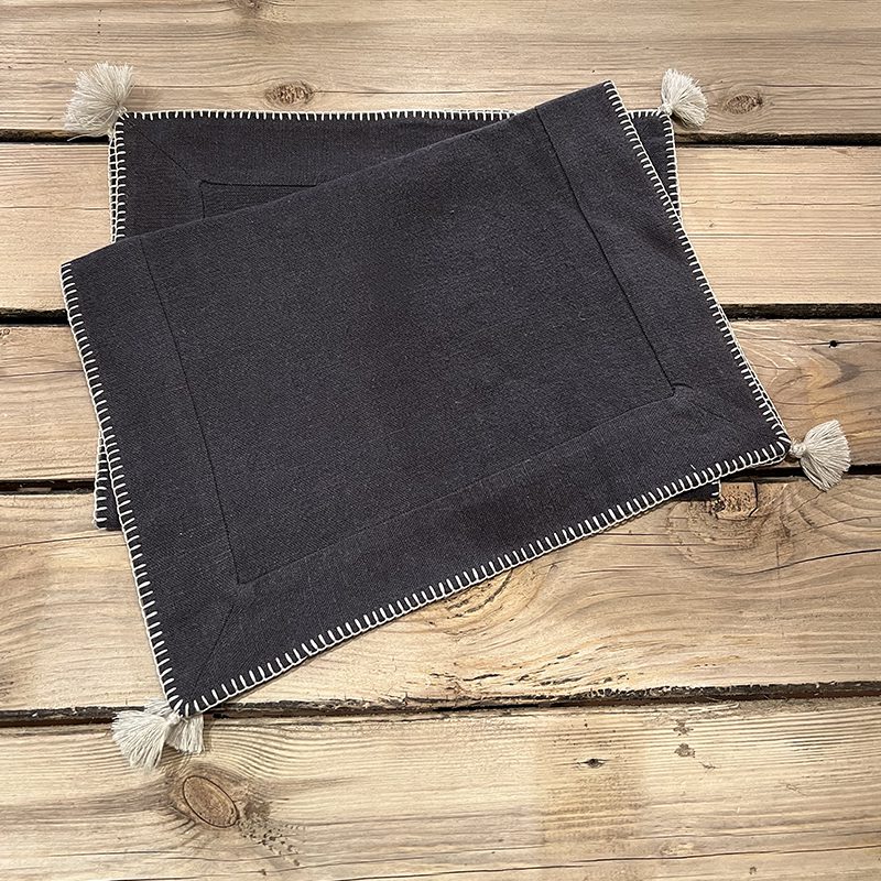 Tibladin Graa Daekkeservietter 1.jpg Sets de table marocains gris anthracite brodés à la main avec bordure blanche et pompons blancs, sur une table en bois