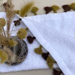 Petite serviette blanche tissée main avec pompons couleur ocre, avec une plante dessus