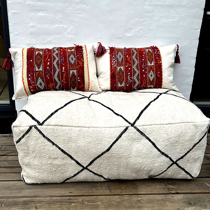 Tibladin Marokkanske Puder Boho 1.jpg Handgeweven vintage kelim boho kussenhoezen in rood en beige met Marokkaans patroon met kwastjes aan de randen staande op een poef